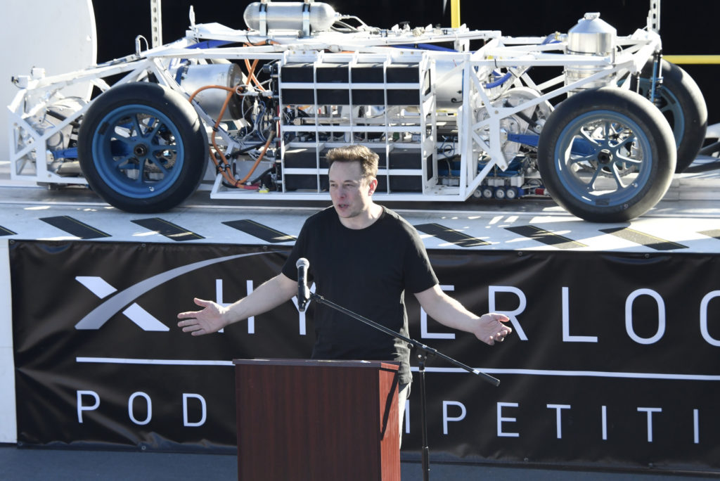 Elon Musk speaking at the SpaceX hyperloop competition in 2017 wearing a black shirt and standing in front of a hyperloop train 
