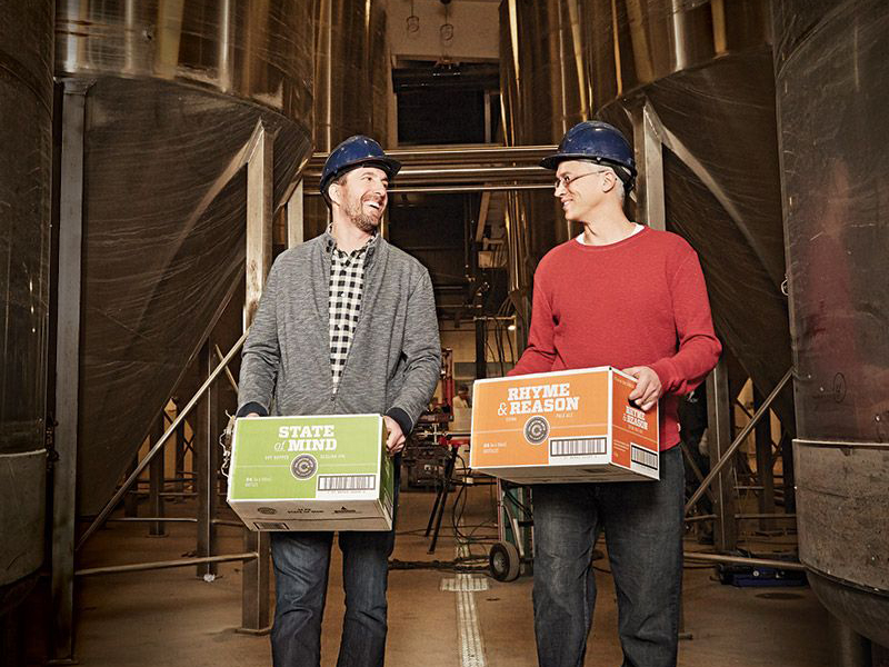 Two men in hard hats walk through a brewery while carrying boxes at Collective Arts Brewing 