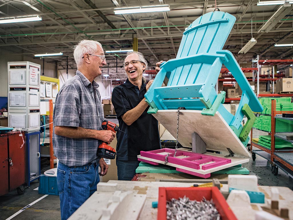 Two men laugh together while working on a Muskoka chair in a factory.