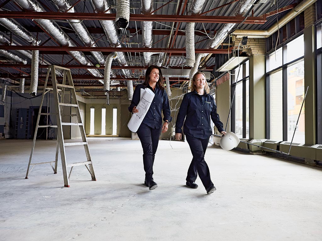 Two women walk through a work site. One holds a roll of paper blueprints and the other holds a hard hat, depicting top canadian entrepreneurs