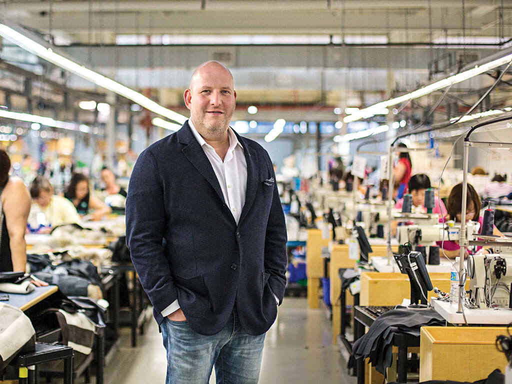 A man in a blazer and jeans stands in the aisle of a factory between rows of sewing machine stations.