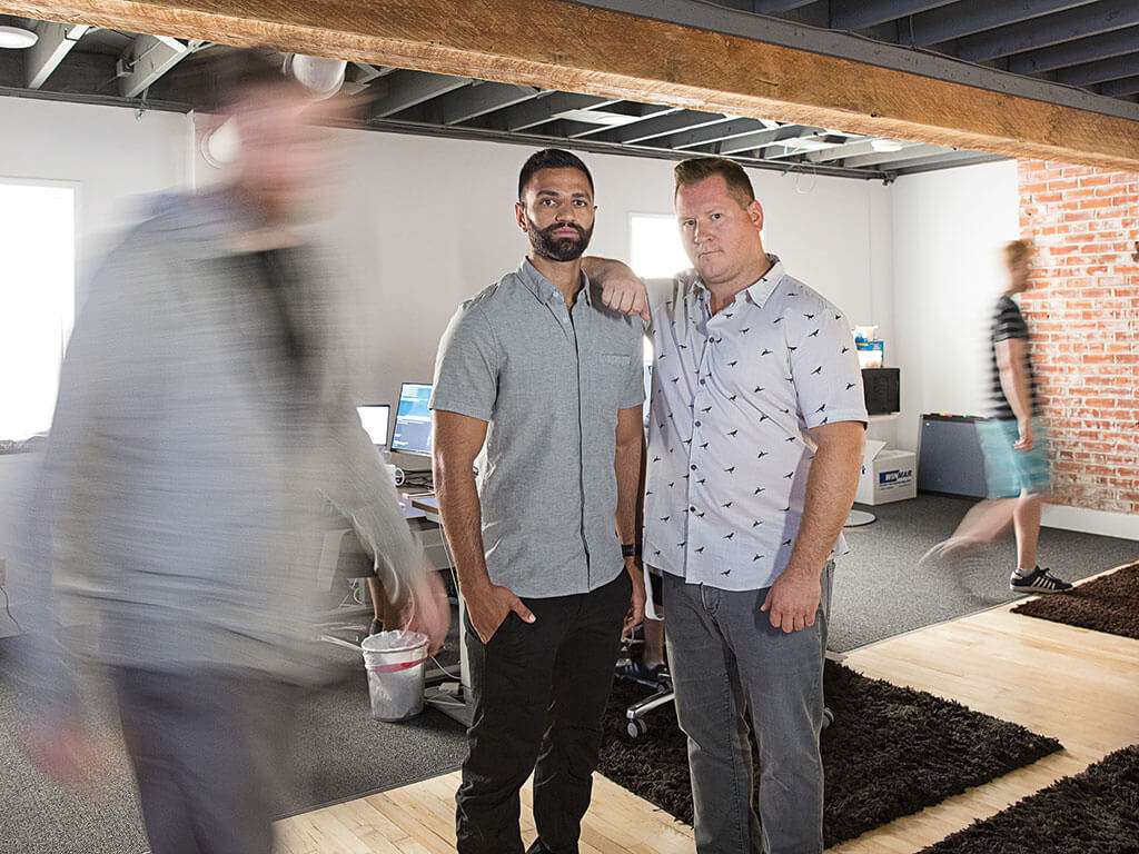 Two top Canadian entrepreneurs in short sleeve button ups pose together for a photo in an office while motion blurred people walk around them.