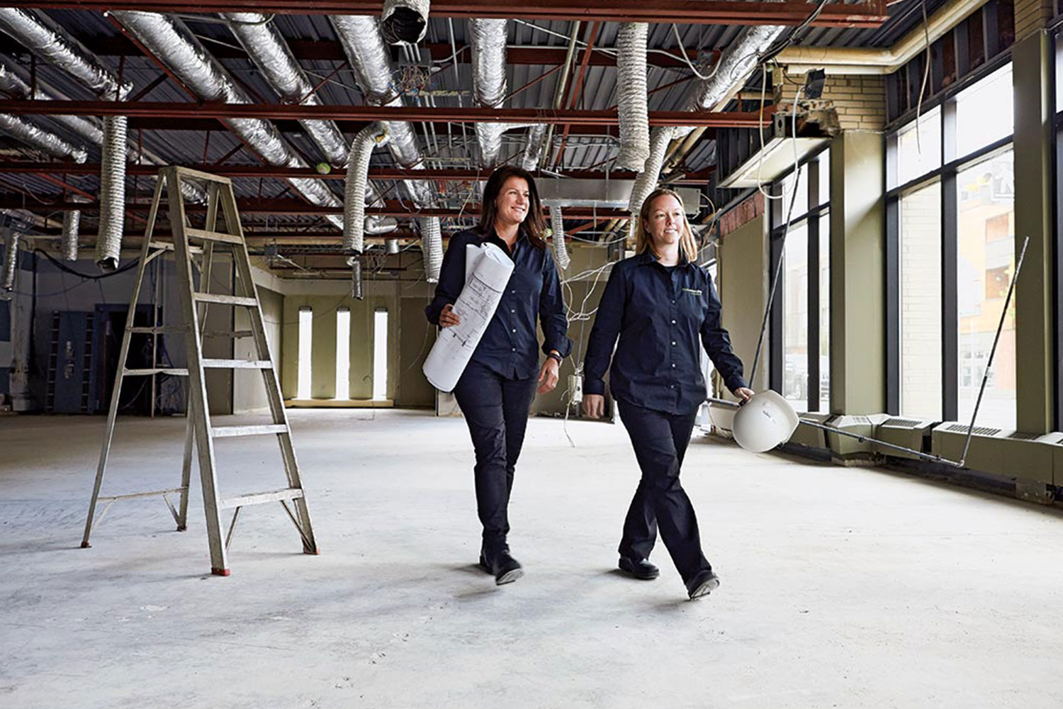 Two women walk through a work site. One holds a roll of paper blueprints and the other holds a hard hat.