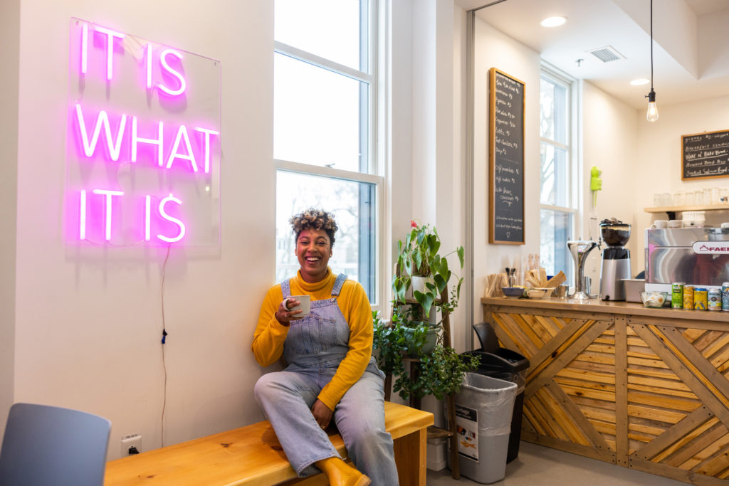 A woman sitting on a bench wearing blue overalls holds a coffee and smiles inside a small cafe 