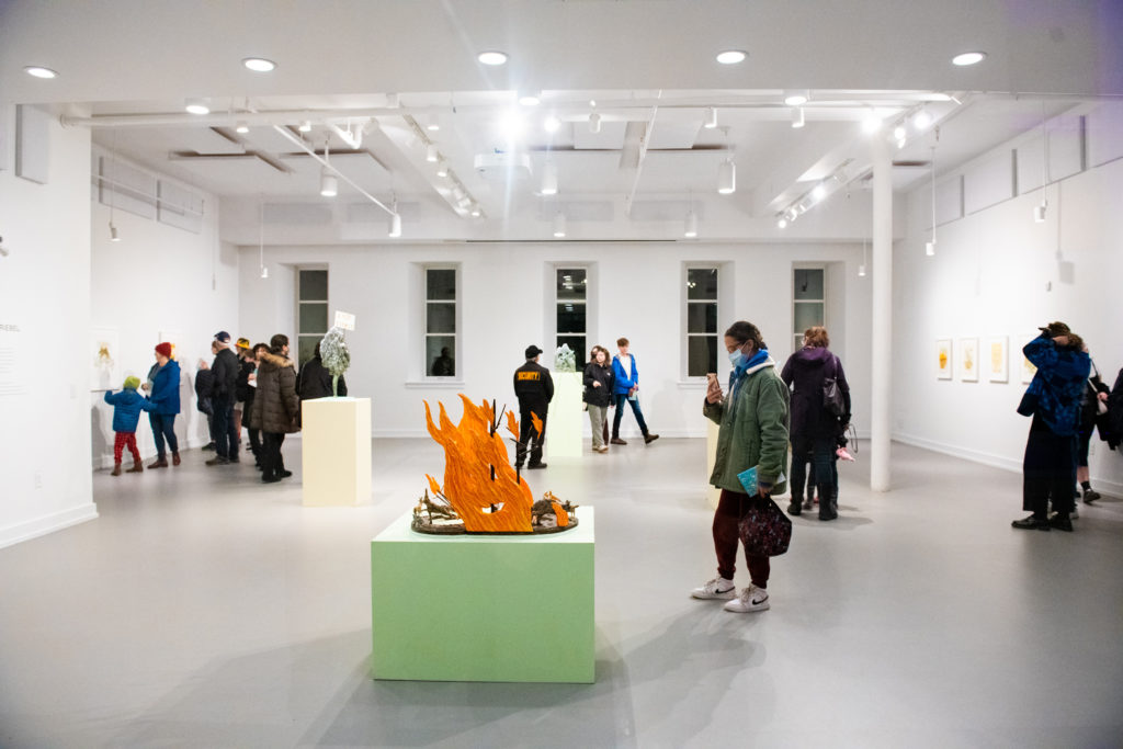 People looking at art inside a white art gallery with a green and orange fire installation in the centre of the white room 
