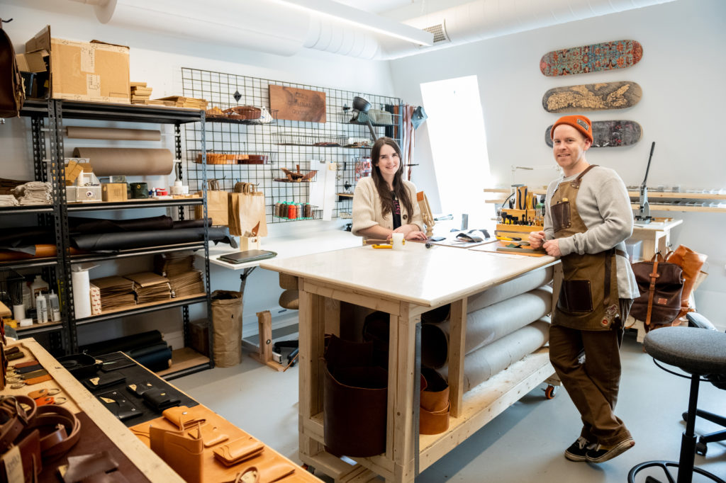 Two people standing inside a leather-making studio. One man is wearing an orange hat and leans against the table. The other, a woman, is smiling with her hands crossed on the table 