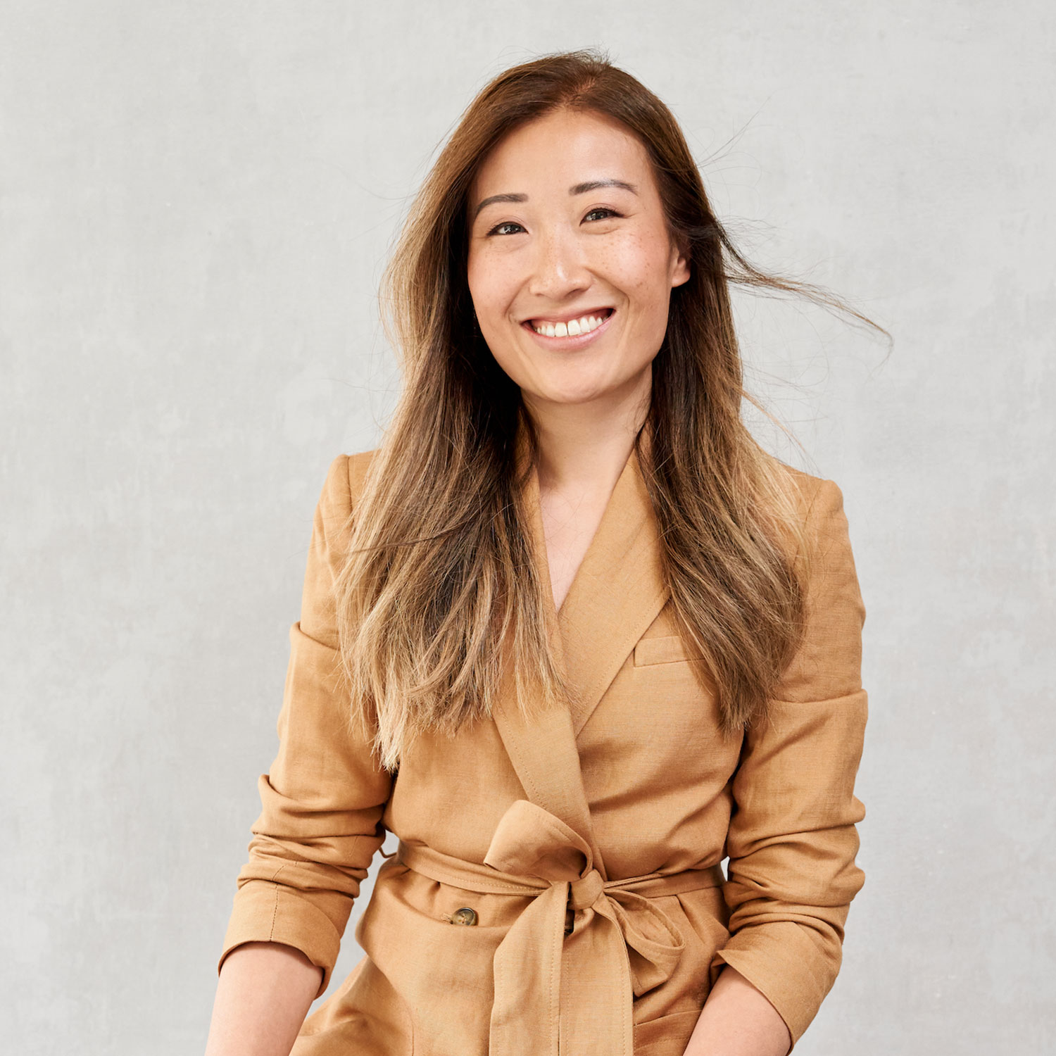 A portrait of Kathy Tran-Riese. She sits in front of a greay background wearing a camel blazer with a wrap closure.