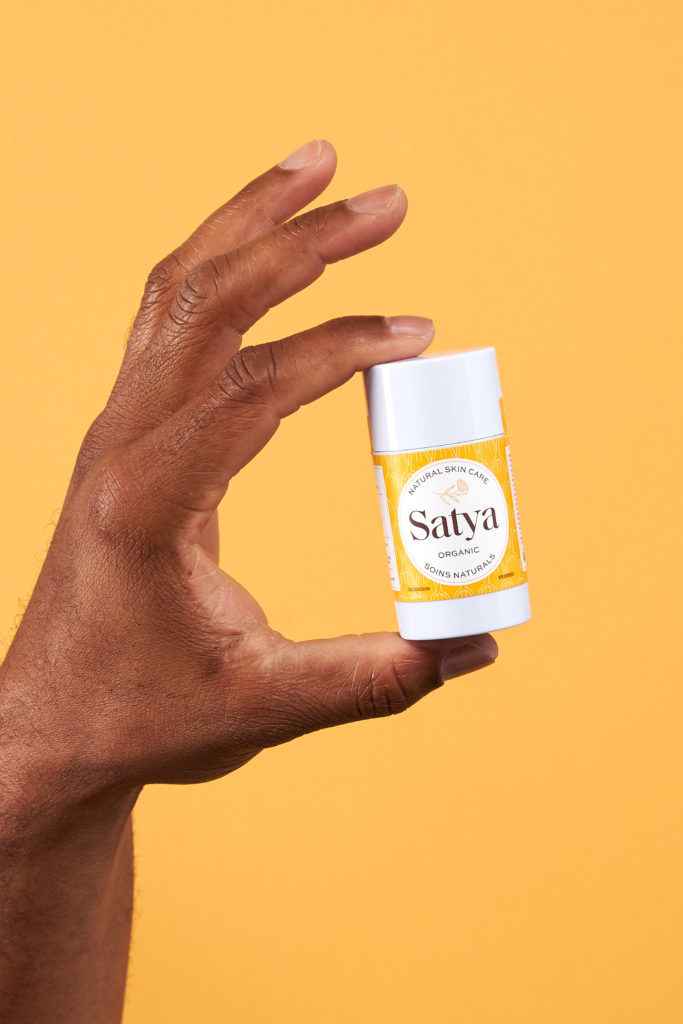 A black hand holding a white container of Satya Organics cream on a yellow background