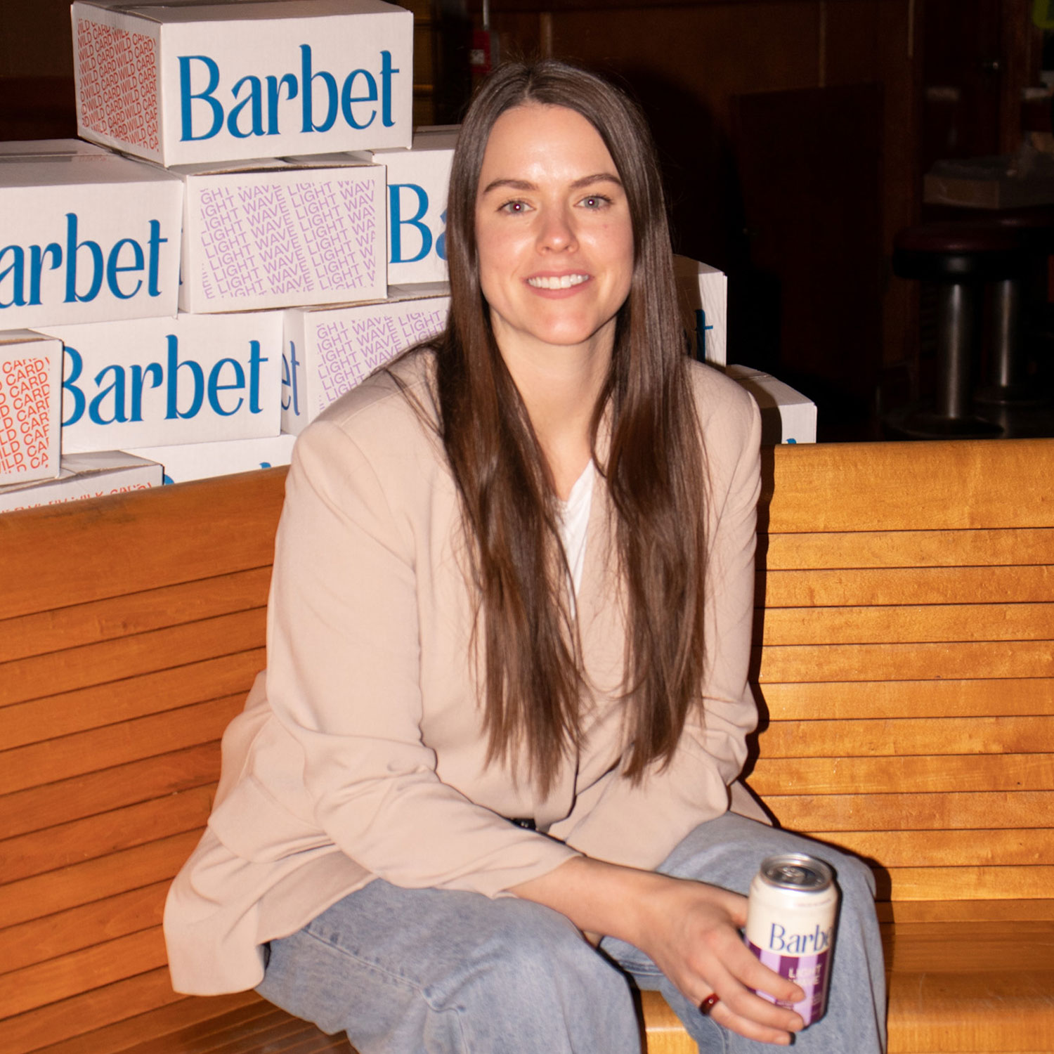 A woman sits on a bench in front of boxes of product while holding a can in her hand. she's wearing a pinkish-beige blazer and light wash jeans