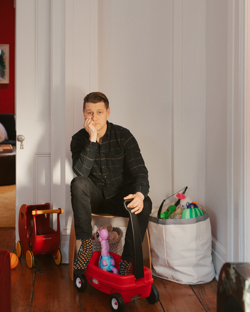 Actor Ben McKenzie sitting on a stool wearing polka dot socks near scattered toys 