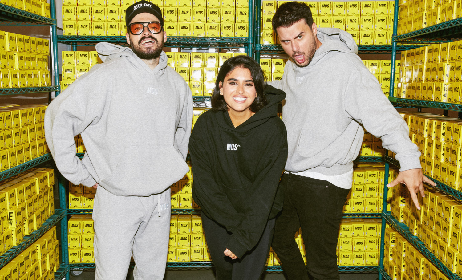 Three people stand before shelves full of product boxes. The two men wear grey branded hoodies while the woman in the middle wears a similar hoodie in black.