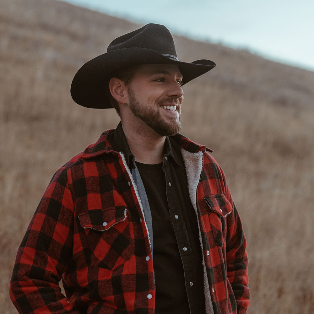 Brett Kissel stands in front of a grassy hill wearing a cowboy hat and a red and black checked jacket