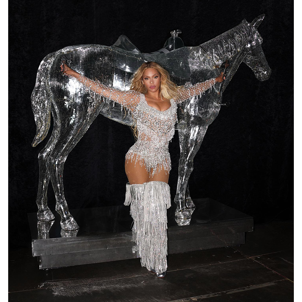 Beyoncé in a shining silver outfit standing in front of a horse statue that is covered in little mirrors like a disco ball.