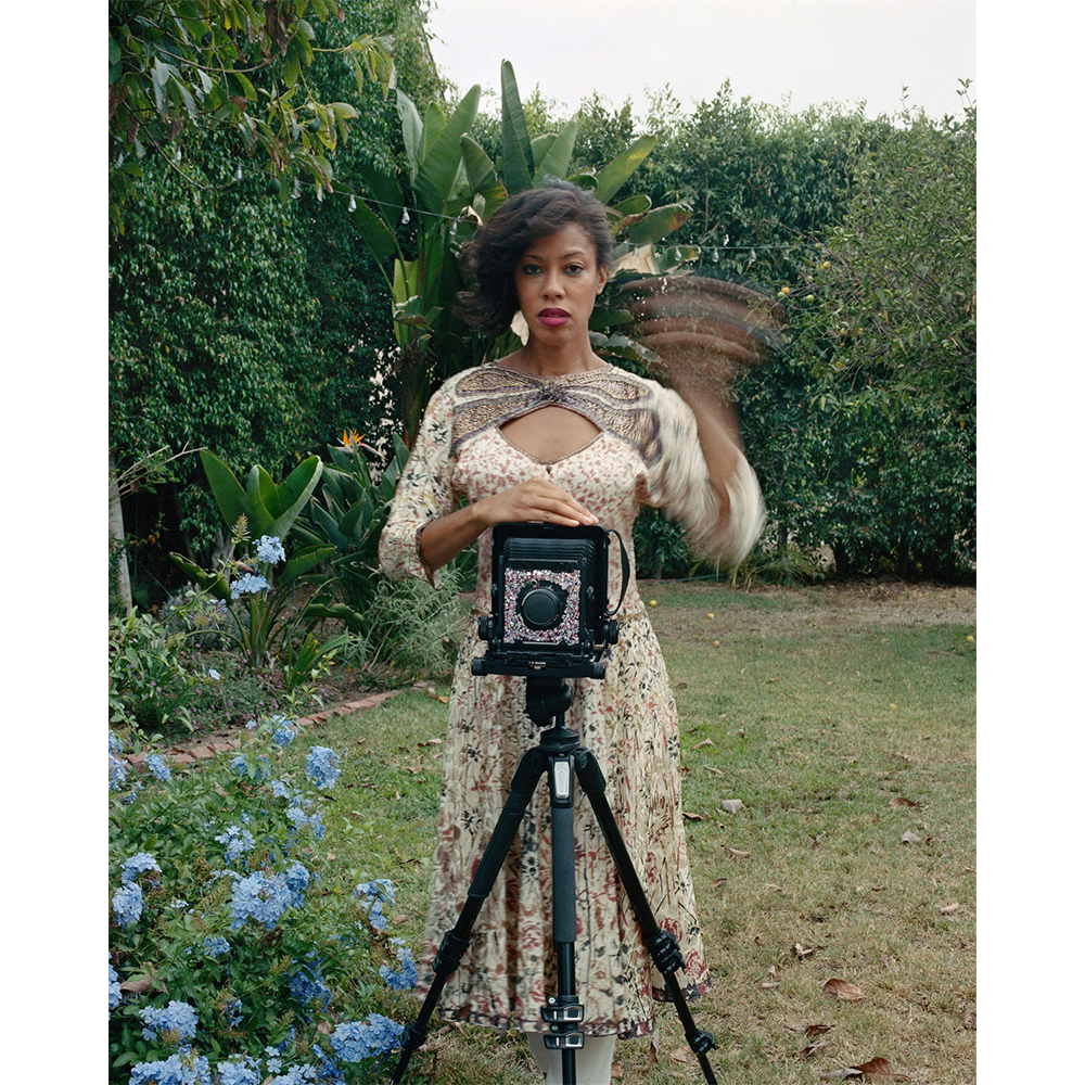 a self portrait photograph of artist Deana Lawson standing in a garden behind and old timey camera on a tripod. Her hand is blurry from waving as the photo was taken.