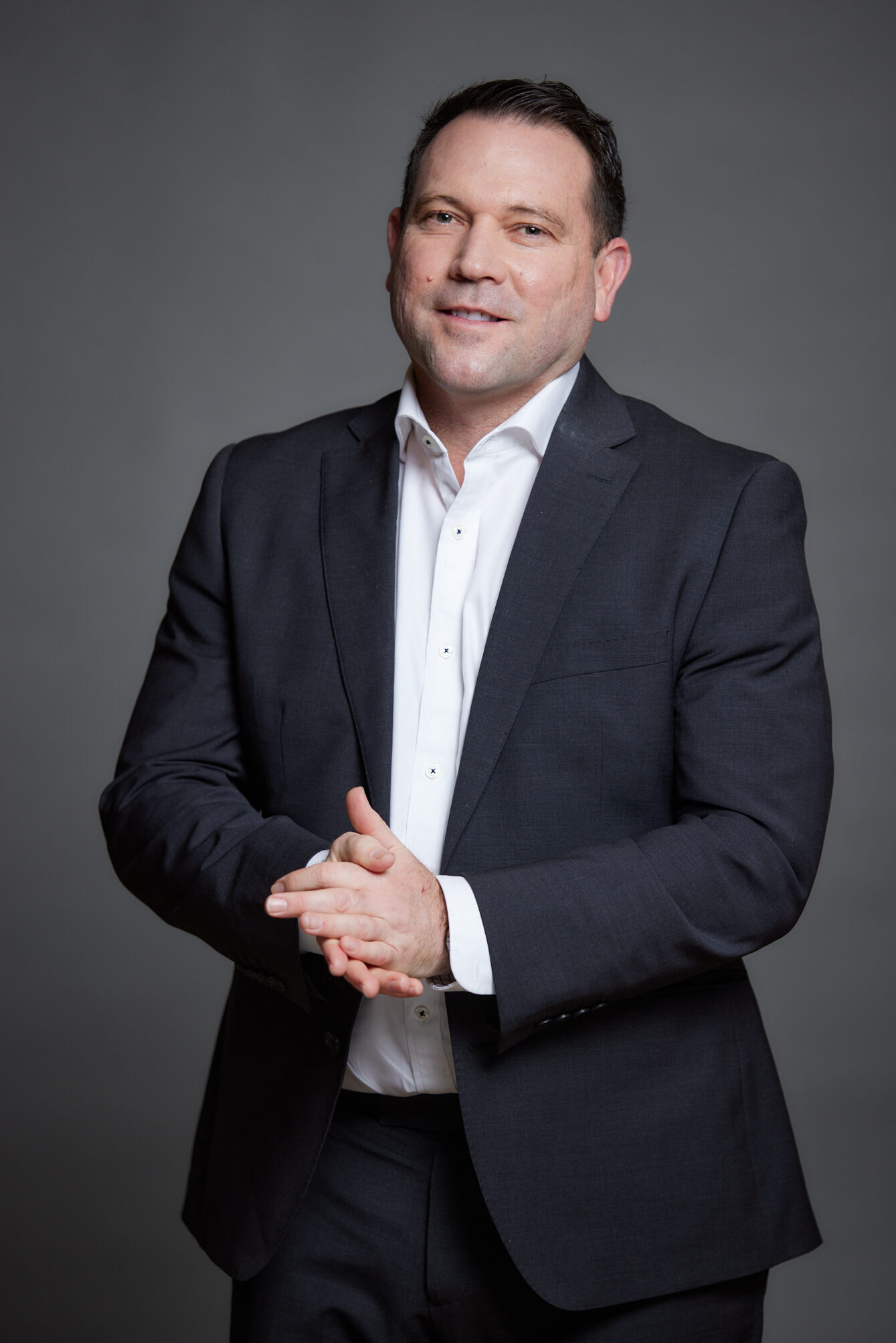 Man in a black suit and white shirt standing with hands clasped against a gray background.