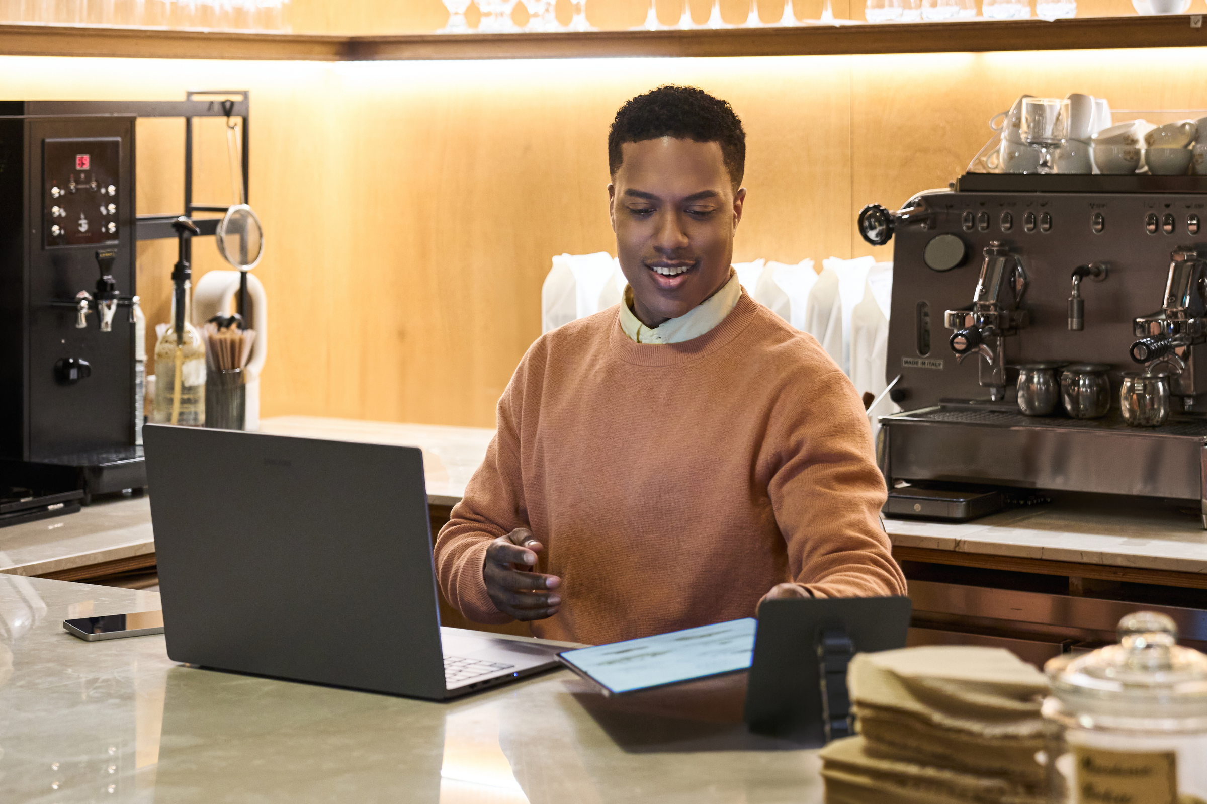 Man wearing a brown sweater and face mask under his chin working on a laptop and tablet at a countertop.