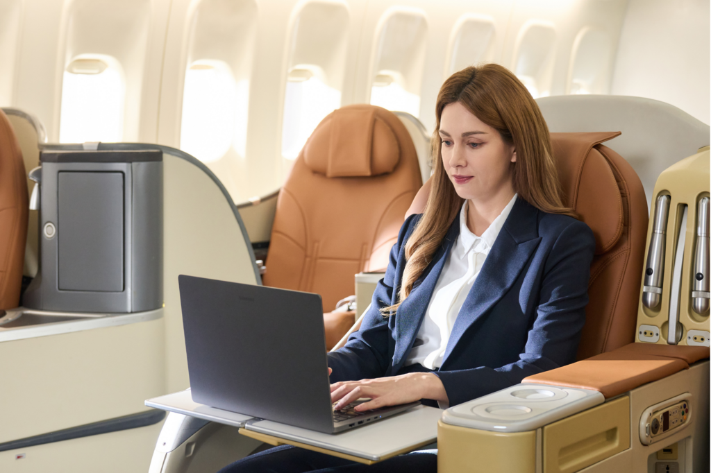 Woman in business attire using laptop while seated in a luxury airplane cabin seat with tan leather upholstery.