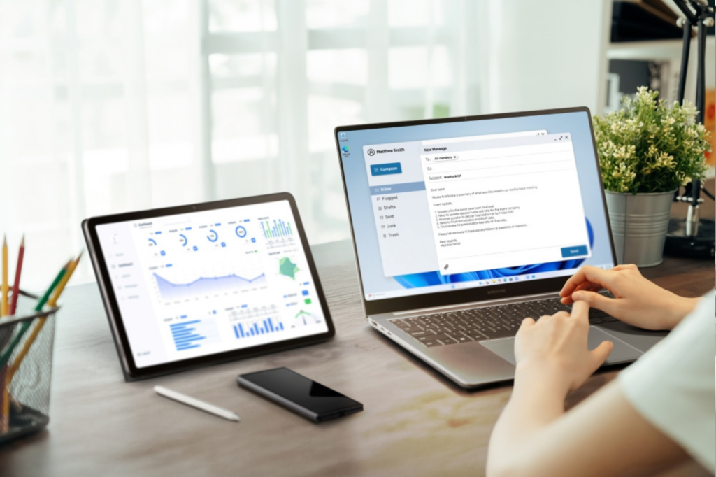 Person using a laptop and tablet with charts and an email draft on a wooden desk with office supplies nearby.