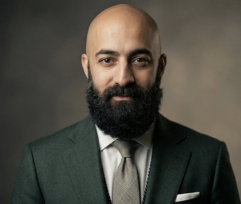 Bearded man with a shaved head wearing a dark green suit and beige tie against a neutral background.