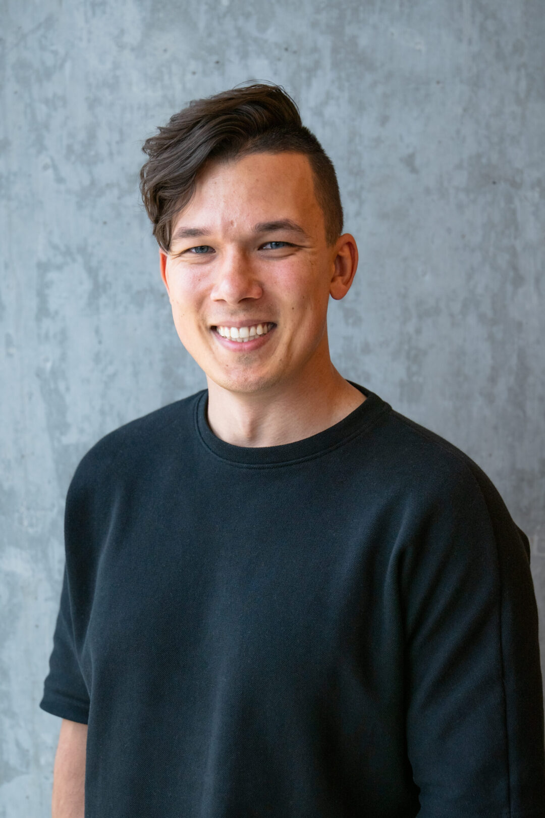 Man with short dark hair wearing a black long-sleeve shirt, smiling against a gray textured wall background.