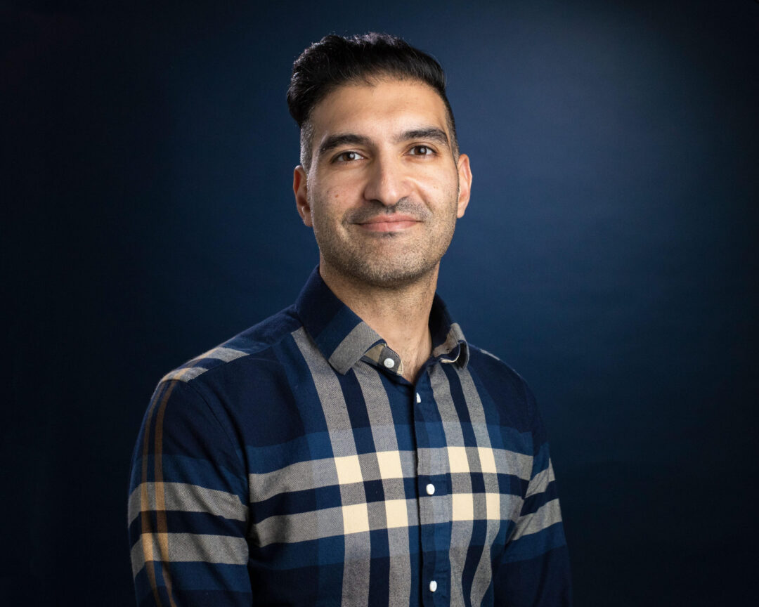 Man with short dark hair wearing a blue and beige plaid shirt, smiling against a dark blue background.