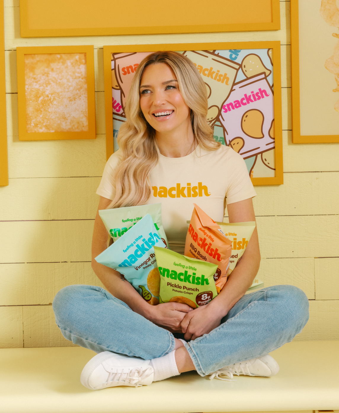 Young woman sitting cross-legged on a yellow bench holding bags of snacks under a sign that reads "feeling a little snackish".